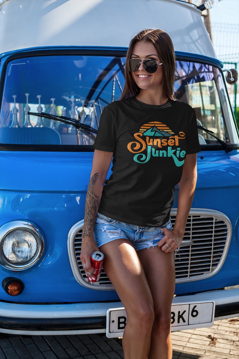 May include: A woman wearing a black t-shirt with a sunset graphic and the text "Sunset Junkie" in a retro font. She is standing in front of a blue van and holding a can of soda.