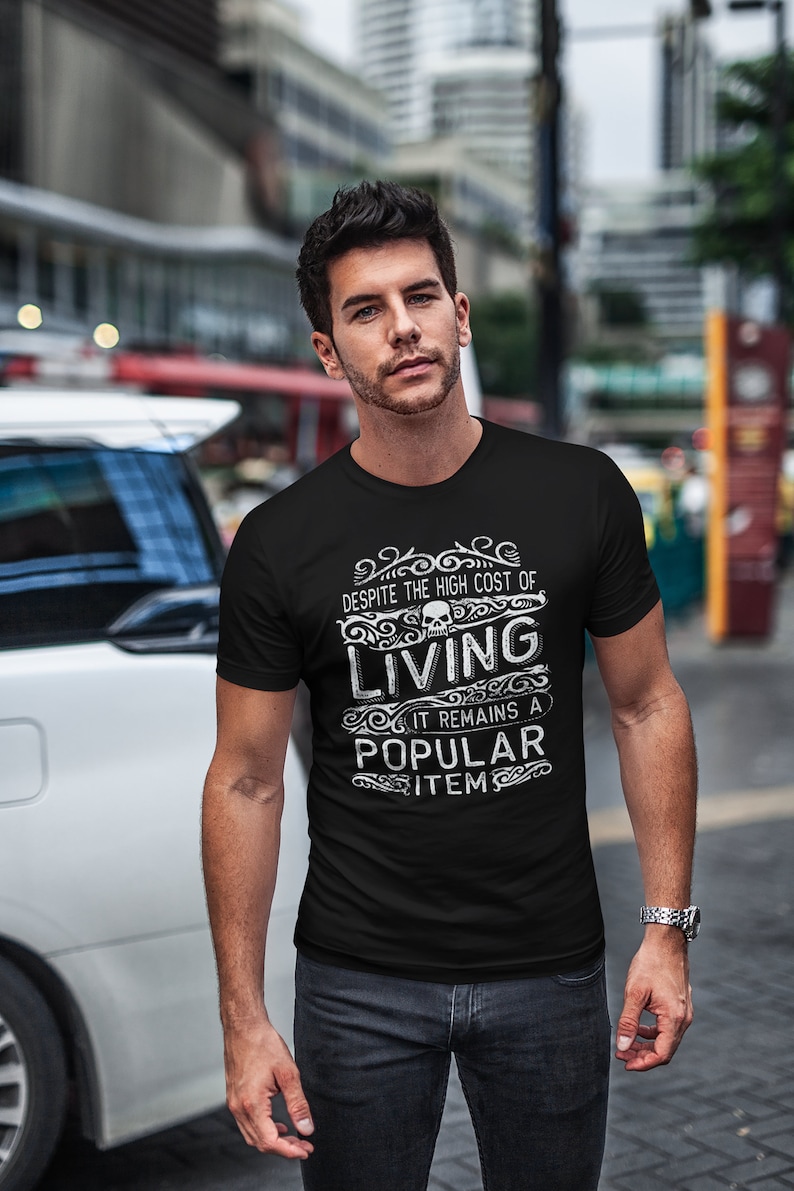May include: A black t-shirt with a white graphic print that says "Despite the high cost of living, it remains a popular item." The graphic also includes a skull and ornate design.