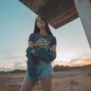 May include: A woman wearing a dark blue t-shirt with a sunset graphic and the text "Sunset Junkie" in a retro font. She is also wearing denim shorts and a denim jacket.