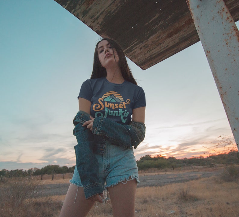 May include: A woman wearing a dark blue t-shirt with a sunset graphic and the text "Sunset Junkie" in a retro font. She is also wearing denim shorts and a denim jacket.