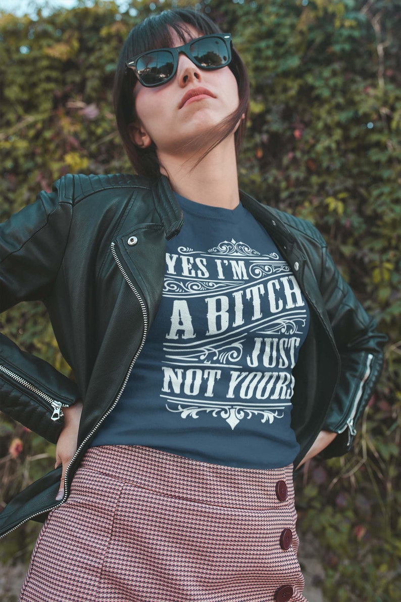 May include: A woman wearing a dark blue t-shirt with the text "Yes I'm a Bitch, Just Not Yours" in white lettering. She is wearing a black leather jacket and a pink and white plaid skirt.