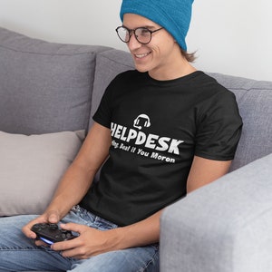 K&ouml;nnte beinhalten: Eine Person tr&auml;gt ein schwarzes T-Shirt mit dem Text "HELPDESK" und einem Headset-Symbol. Der Text unter dem Logo lautet "Sing Beat if You Moron". Die Person tr&auml;gt eine blaue M&uuml;tze und eine Brille und h&auml;lt einen Videospiel-Controller.