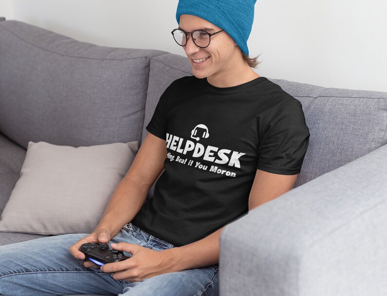 K&ouml;nnte beinhalten: Eine Person tr&auml;gt ein schwarzes T-Shirt mit dem Text "HELPDESK" und einem Headset-Symbol. Der Text unter dem Logo lautet "Sing Beat if You Moron". Die Person tr&auml;gt eine blaue M&uuml;tze und eine Brille und h&auml;lt einen Videospiel-Controller.