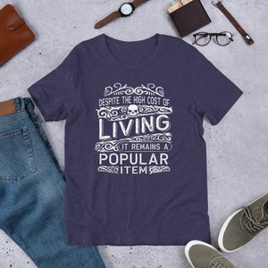 May include: A dark blue t-shirt with a white graphic design that says "Despite the high cost of living it remains a popular item" with a skull graphic in the middle of the text.