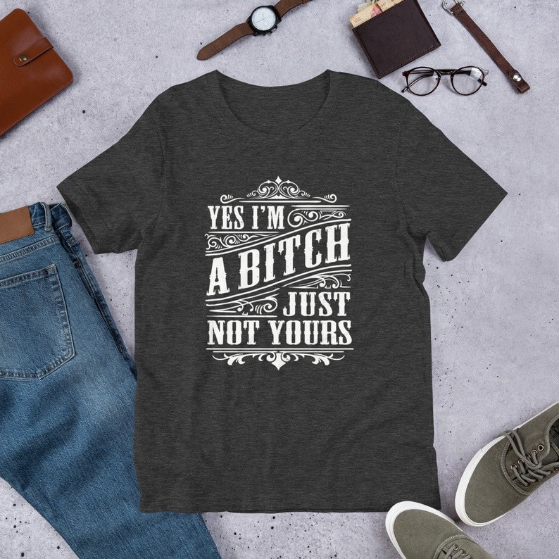 May include: Dark heather grey t-shirt with a white graphic that reads "Yes I'm a Bitch Just Not Yours" in a vintage style font.