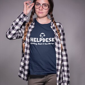 K&ouml;nnte beinhalten: Eine Frau tr&auml;gt ein marineblaues T-Shirt mit einer wei&szlig;en Grafik eines Headsets und dem Text "Helpdesk Kidding. Beat if You Moron." Sie tr&auml;gt auch ein schwarz-wei&szlig; kariertes Hemd und eine braune Hose.