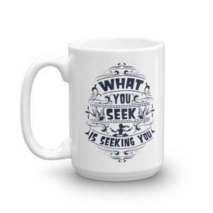 May include: White ceramic coffee mug with a blue design that says "What you seek is seeking you." The design is in a circular frame with a decorative border.