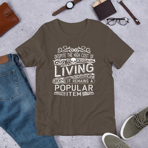 May include: A brown t-shirt with a white graphic design that says "Despite the high cost of living, it remains a popular item." The graphic design includes a skull and ornate scrollwork.