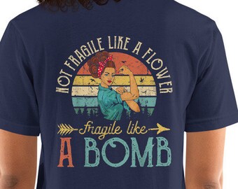 May include: Navy blue t-shirt with a vintage-style graphic of a woman with her fist raised. The graphic says "Not Fragile Like A Flower - Fragile Like A Bomb".