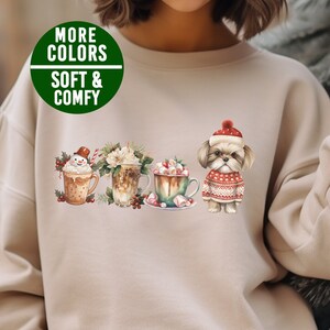 May include: Beige sweatshirt with a Christmas-themed design featuring a snowman, a cup of hot chocolate, a cup of coffee, and a Shih Tzu dog wearing a Santa hat. The text "More Colors Soft & Comfy" is printed on the top left corner of the sweatshirt.