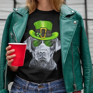 以下が含まれることがあります： 黒いTシャツに、緑の妖精の帽子とサングラスをかけた黒いラブラドール犬のグラフィックがプリントされています。犬はカメラを見つめています。