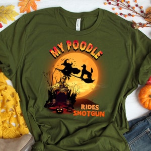 May include: A green t-shirt with a graphic of a witch and a poodle flying on a broomstick in front of a full moon and a haunted house. The text "My Poodle Rides Shotgun" is printed on the shirt.