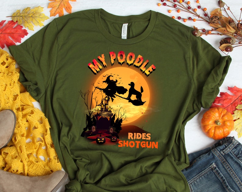 May include: A green t-shirt with a graphic of a witch and a poodle flying on a broomstick in front of a full moon and a haunted house. The text "My Poodle Rides Shotgun" is printed on the shirt.