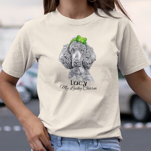 Pu&ograve; includere: Una maglietta bianca con un'illustrazione in bianco e nero di un barboncino che indossa un cerchietto verde con un quadrifoglio. Il testo "Lucy My Lucky Charm" &egrave; stampato sotto l'immagine.