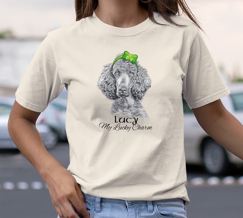 Pu&ograve; includere: Una maglietta bianca con un'illustrazione in bianco e nero di un barboncino che indossa un cerchietto verde con un quadrifoglio. Il testo "Lucy My Lucky Charm" &egrave; stampato sotto l'immagine.