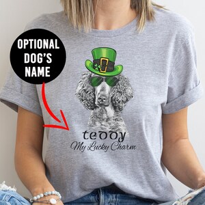 Pu&ograve; includere: Una maglietta grigia con un'immagine in bianco e nero di un barboncino che indossa un cappello verde da folletto e occhiali da sole. Il testo "teddy My Lucky Charm" &egrave; stampato sotto l'immagine.