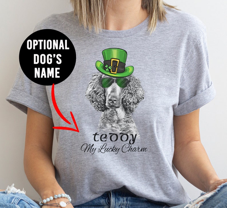 Pu&ograve; includere: Una maglietta grigia con un'immagine in bianco e nero di un barboncino che indossa un cappello verde da folletto e occhiali da sole. Il testo "teddy My Lucky Charm" &egrave; stampato sotto l'immagine.