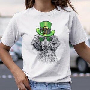 Pu&ograve; includere: Maglietta bianca con una grafica di un cane che indossa un cappello verde da folletto e occhiali da sole. Il cane &egrave; un barboncino grigio.