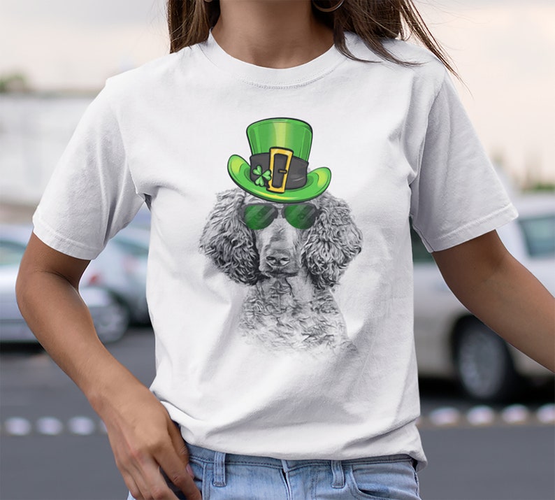 Pu&ograve; includere: Maglietta bianca con una grafica di un cane che indossa un cappello verde da folletto e occhiali da sole. Il cane &egrave; un barboncino grigio.