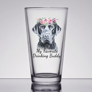 May include: A clear glass pint glass with a black labrador retriever wearing a pink and white floral crown. The text "My Favorite Drinking Buddy" is printed on the glass.