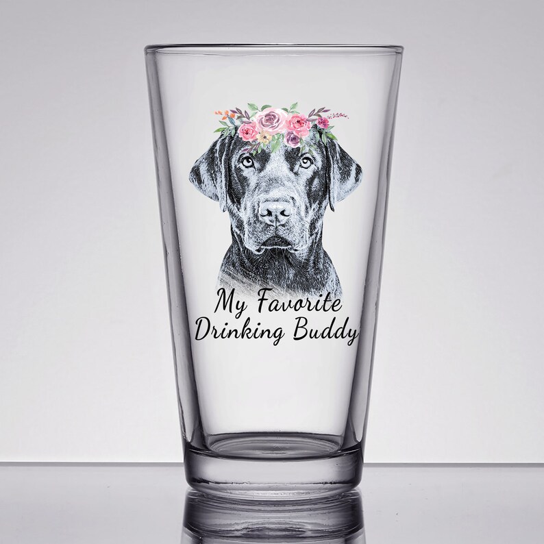 May include: A clear glass pint glass with a black labrador retriever wearing a pink and white floral crown. The text "My Favorite Drinking Buddy" is printed on the glass.