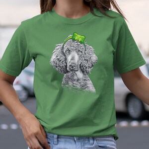Pu&ograve; includere: Una maglietta verde con un'illustrazione in bianco e nero di un barboncino che indossa un cerchietto verde a forma di trifoglio.