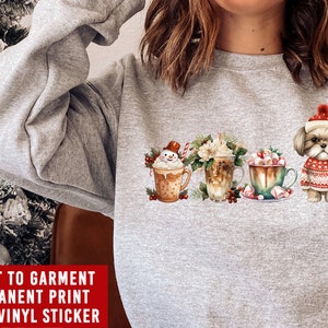 May include: A gray sweatshirt with a Christmas-themed design featuring a snowman, a cup of coffee, a cup of hot chocolate, and a dog wearing a Santa hat. The text "DIRECT TO GARMENT PERMANENT PRINT NOT A VINYL STICKER" is printed on the bottom of the sweatshirt.