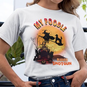 May include: White t-shirt with a graphic of a poodle riding with a witch on a broomstick in front of a full moon. The text "My Poodle Rides Shotgun" is printed on the shirt.