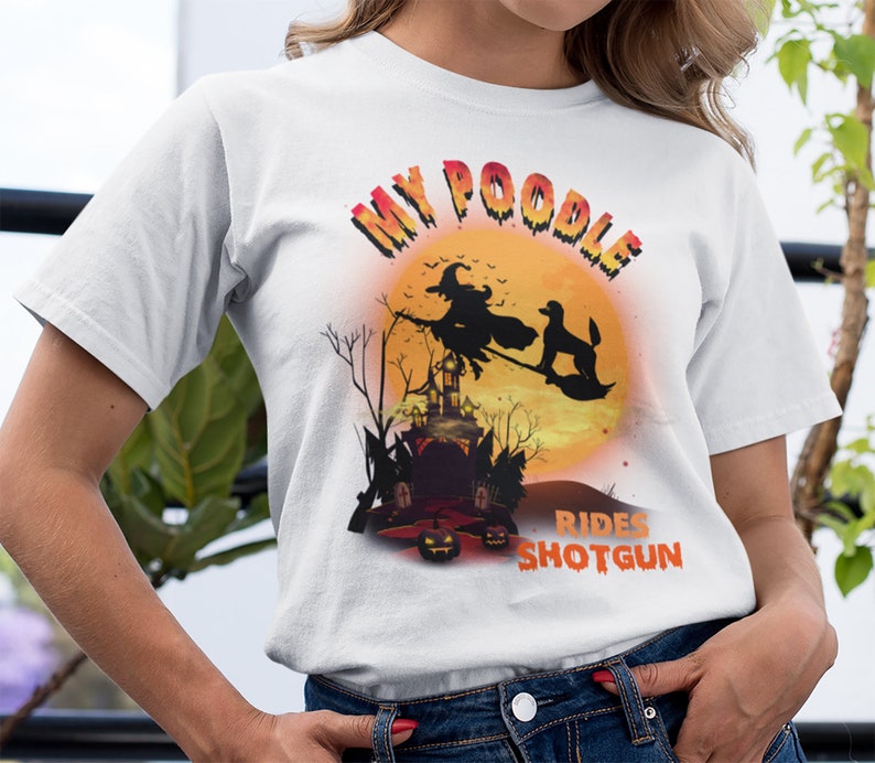 May include: White t-shirt with a graphic of a poodle riding with a witch on a broomstick in front of a full moon. The text "My Poodle Rides Shotgun" is printed on the shirt.
