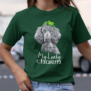 Pu&ograve; includere: Una maglietta verde con un'illustrazione in bianco e nero di un barboncino che indossa un cerchietto con un trifoglio. Il testo "My Lucky Charm" &egrave; stampato sotto il barboncino.
