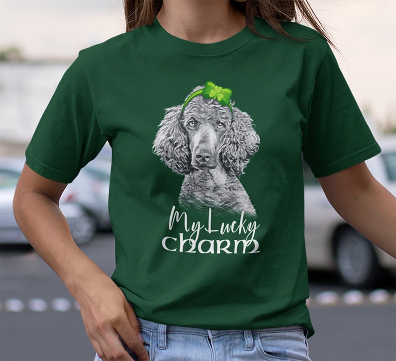 Pu&ograve; includere: Una maglietta verde con un'illustrazione in bianco e nero di un barboncino che indossa un cerchietto con un trifoglio. Il testo "My Lucky Charm" &egrave; stampato sotto il barboncino.