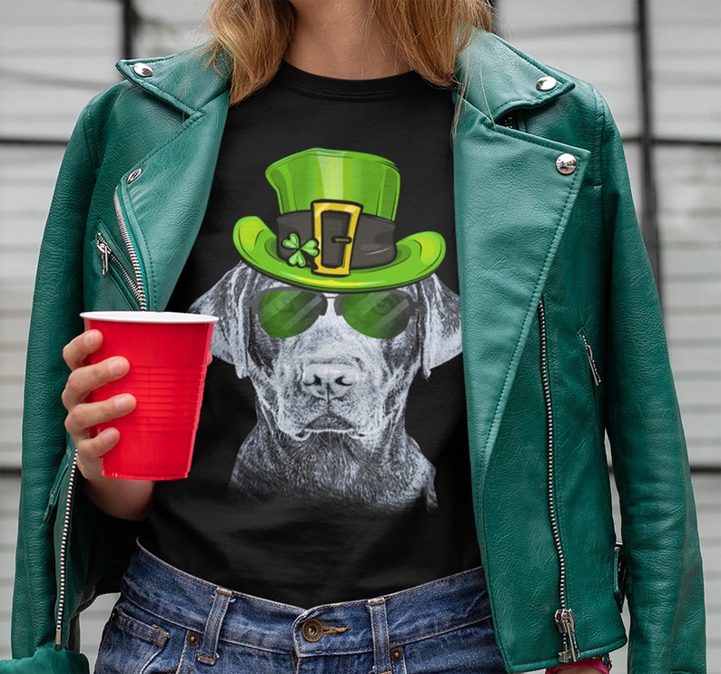 以下が含まれることがあります： 黒いTシャツに、緑の妖精の帽子とサングラスをかけた黒いラブラドール犬のグラフィックがプリントされています。犬はカメラを見つめています。
