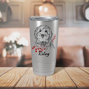 May include: A silver travel mug with a detailed grayscale portrait of a dog, red heart accents, and the name "Riley" in black script. The mug has a silver lid and is set on a wooden surface.
