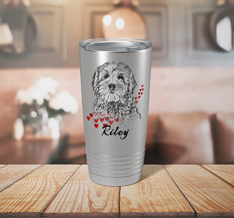 May include: A silver travel mug with a detailed grayscale portrait of a dog, red heart accents, and the name "Riley" in black script. The mug has a silver lid and is set on a wooden surface.