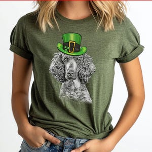 Pu&ograve; includere: Una maglietta verde con un'illustrazione in bianco e nero di un barboncino che indossa un cappello verde da folletto. Il cappello ha una fibbia dorata e un trifoglio.