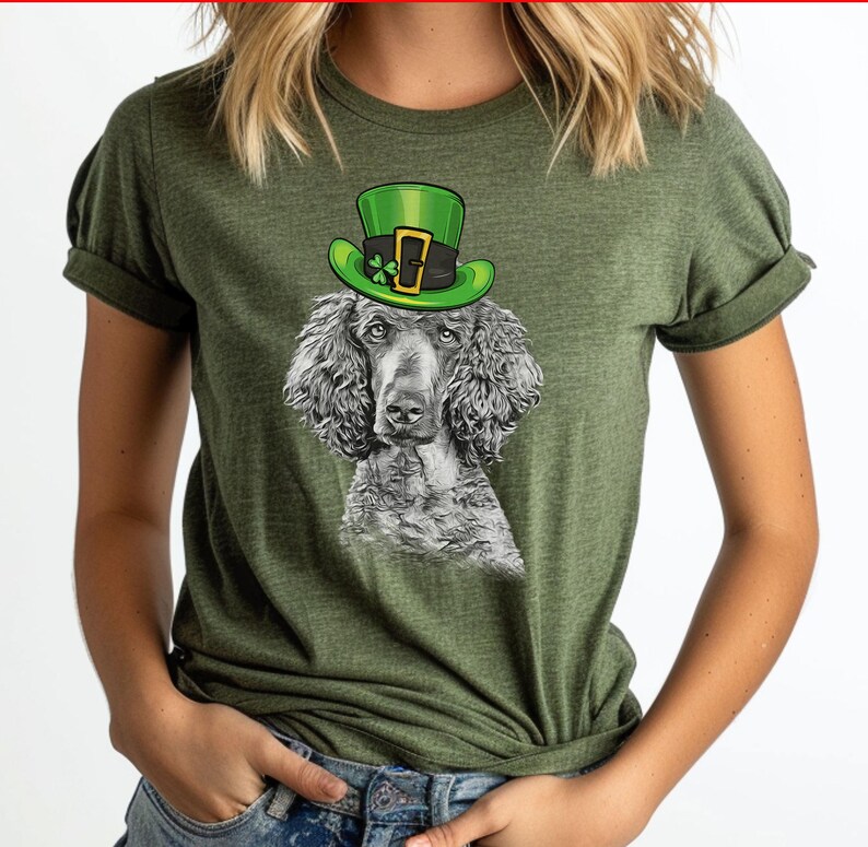 Pu&ograve; includere: Una maglietta verde con un'illustrazione in bianco e nero di un barboncino che indossa un cappello verde da folletto. Il cappello ha una fibbia dorata e un trifoglio.