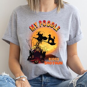 May include: Gray t-shirt with a graphic of a poodle riding a broomstick with a witch in front of a full moon and a haunted house. The text reads "My Poodle Rides Shotgun".