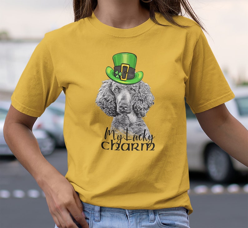 Pu&ograve; includere: Una maglietta gialla con un barboncino grigio che indossa un cappello verde da folletto. Il testo "My Lucky Charm" &egrave; stampato sulla maglietta.