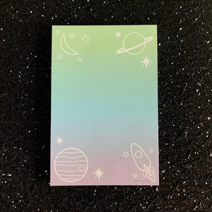 May include: A pastel gradient notepad with a rocket, planets, stars, and a crescent moon. The notepad has the text "Fortuna Pins" at the bottom.