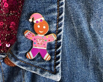 May include: A pink gingerbread man enamel pin with a red heart on a sweater, wearing a pink hat with a white pom-pom. The pin is attached to a blue denim jacket.