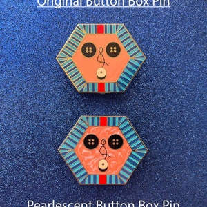 May include: Two hexagonal pins with a face design. The top pin is labeled "Original Button Box Pin." The bottom pin is labeled "Pearlescent Button Box Pin." Each pin has a peach-colored face, black button eyes, and a red and blue striped border.