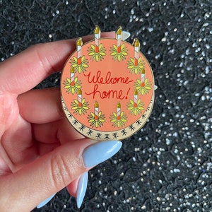 May include: A round enamel pin with a peach-colored background featuring the text "Welcome home!" in red script. The design includes a circular border of yellow flowers with tall, striped candles topped with gold glitter flames.