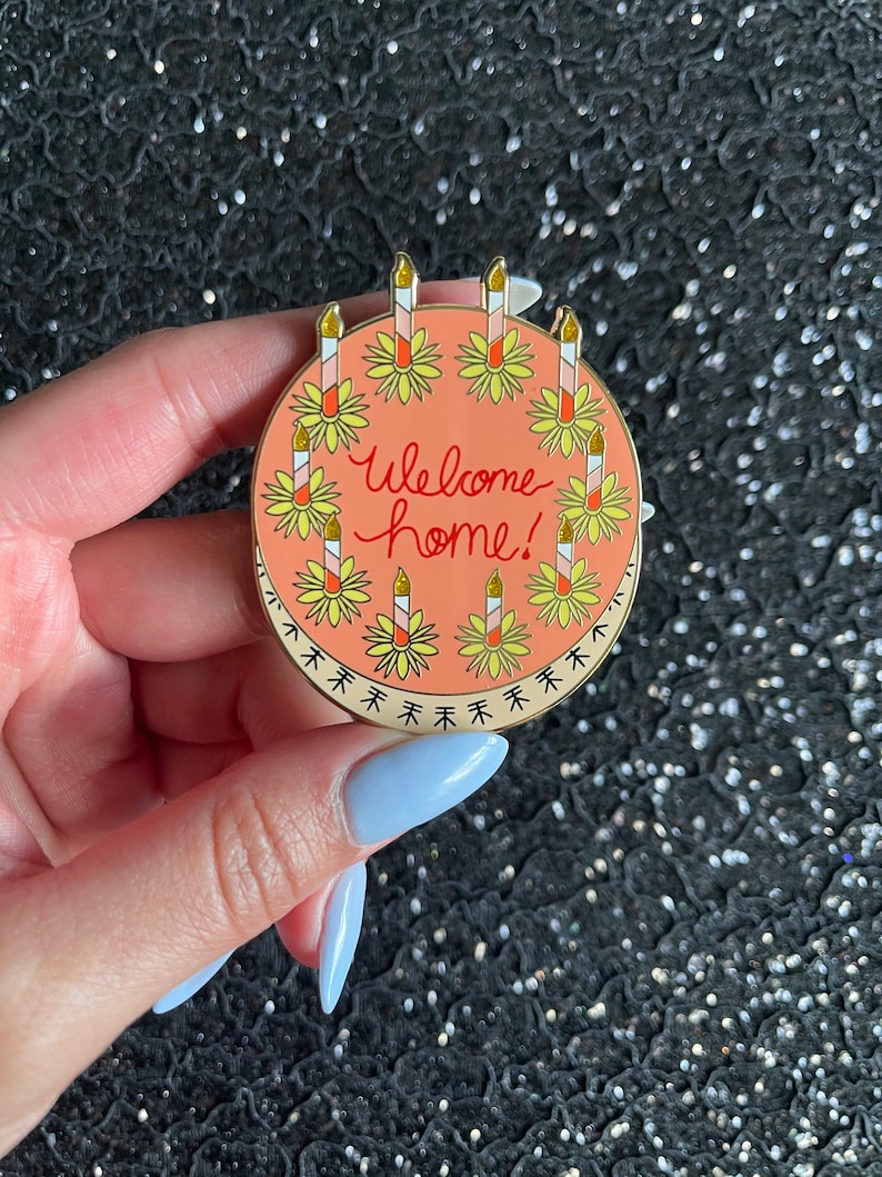 May include: A round enamel pin with a peach-colored background featuring the text "Welcome home!" in red script. The design includes a circular border of yellow flowers with tall, striped candles topped with gold glitter flames.