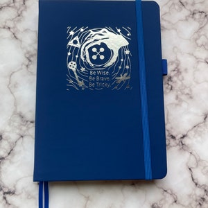 Op de afbeelding: Een donkerblauw notitieboek met een zilveren folieontwerp met een knoop, een sleutel en een libel. Het ontwerp is omringd door sterren en de tekst "Be Wise. Be Brave. Be Tricky."