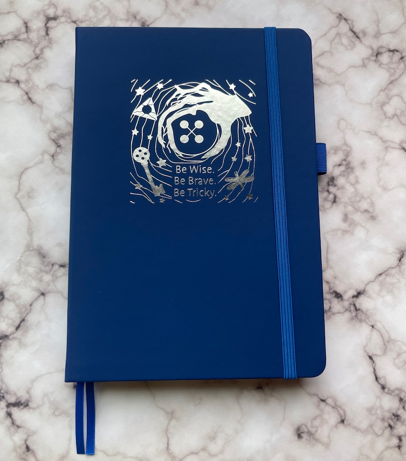 Op de afbeelding: Een donkerblauw notitieboek met een zilveren folieontwerp met een knoop, een sleutel en een libel. Het ontwerp is omringd door sterren en de tekst "Be Wise. Be Brave. Be Tricky."