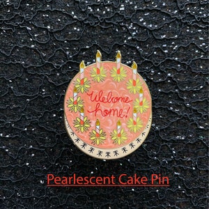May include: A round enamel pin featuring a pearlescent peach-colored cake with the text Welcome home! written in red script. The cake is decorated with eight lit candles, each surrounded by a yellow flower, and a border of small black arrows.
