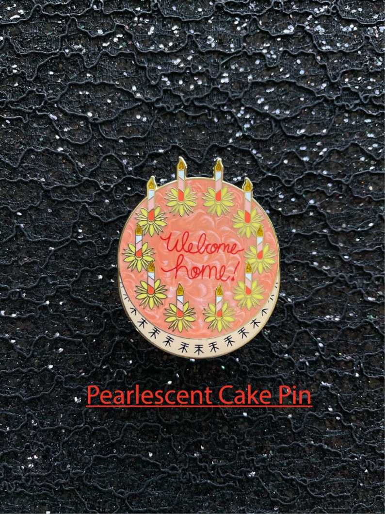 May include: A round enamel pin featuring a pearlescent peach-colored cake with the text Welcome home! written in red script. The cake is decorated with eight lit candles, each surrounded by a yellow flower, and a border of small black arrows.