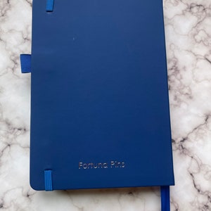 Op de afbeelding: Een donkerblauw notitieboek met een blauw elastiekje en een blauw lint als bladwijzer. Op de voorkant van het notitieboek staat "Fortuna Pins" in goud gegraveerd.