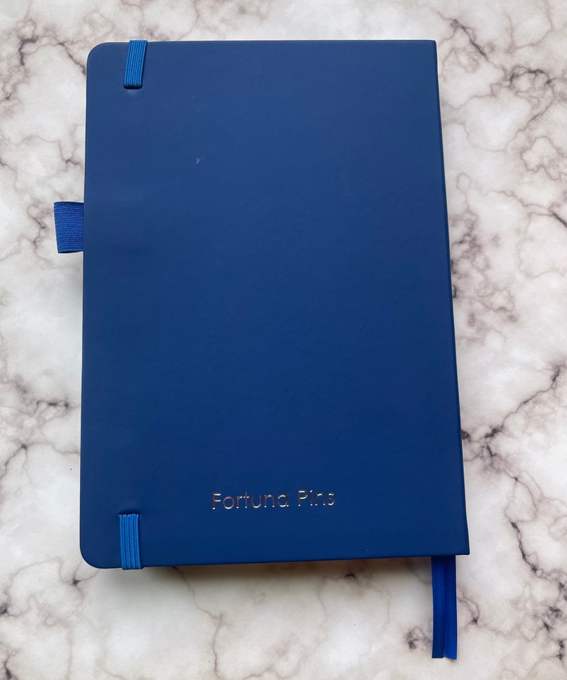 Op de afbeelding: Een donkerblauw notitieboek met een blauw elastiekje en een blauw lint als bladwijzer. Op de voorkant van het notitieboek staat "Fortuna Pins" in goud gegraveerd.