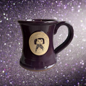 May include: A deep purple ceramic mug with a curved handle. The mug features a circular emblem with a black and white illustration of a character. The mug has a glossy finish and a brown base.